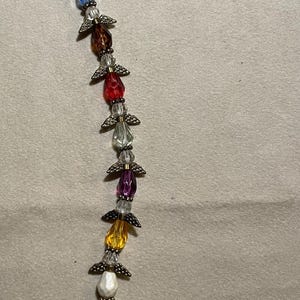 Op de afbeelding: Een kralen engel ornament met een verscheidenheid aan kleurrijke druppelvormige kralen. Elke kraal is geaccentueerd met bronzen metalen vleugels. De kralen zijn onder meer blauw, zwart, rood, paars, geel en roze.