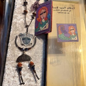 May include: A necklace featuring a silver-toned face pendant with a crescent-shaped base, dangling orange and blue beads, and metallic accents. It is presented in a box with a tag and a clear lid, both displaying the text Tickle Your Soul by Kimberly Wilcox.