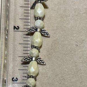 Pode incluir: Uma pulseira de contas de anjo, com aproximadamente 10 cm de comprimento, com contas em forma de lágrima em branco sujo e detalhes de asas de anjo prateadas. A pulseira é medida contra uma régua.