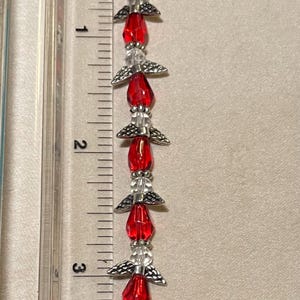 May include: A beaded ornament featuring red, faceted beads and silver-toned angel wing accents. The ornament is approximately 3.5 inches long and is displayed next to an inch ruler.