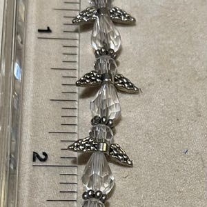 Op de afbeelding: Een heldere kristallen engelarmband, ongeveer 7,6 cm lang, met zilverkleurige metalen vleugels en accenten. De armband wordt naast een liniaal getoond.