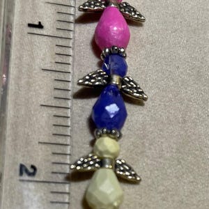 Könnte beinhalten: Ein Armband mit Perlen-Engeln mit weißen, rosa, blauen, gelben und grünen facettierten Perlen. Jede Perle wird durch kleine silberfarbene Metallflügel getrennt. Das Armband ist etwa 5 cm lang.