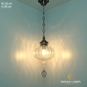 May include: A clear glass pendant light with a ribbed design hangs from a silver chain. The light is suspended from a silver ceiling fixture. The light has a decorative silver ornament hanging from the bottom. The light is on and illuminating the room. The dimensions of the light are 22 cm wide and 83 cm high.