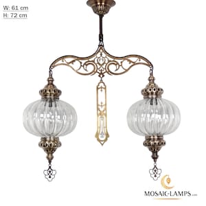 Puede incluir: Una lámpara de araña ornamentada de estilo antiguo con dos globos de vidrio transparente y acanalado. La lámpara tiene un acabado en bronce con filigranas decorativas y un colgante central. Ancho 61 cm, alto 72 cm. El logotipo de MOSAIC-LAMPS.com es visible.