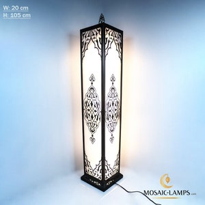 Op de afbeelding: Een hoge zwarte staande lamp met een witte, matglazen kap. De lamp heeft ingewikkelde geometrische ontwerpen in een zwart metalen frame. De lamp is 105 cm hoog en 20 cm breed.