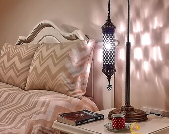 Puede incluir: Una lámpara de mesa de metal negro con una pantalla cilíndrica decorativa y adornada. La lámpara está encendida y proyecta una luz cálida. La lámpara está colocada sobre una mesita de noche blanca junto a una cama con un edredón estampado en beige y blanco. La lámpara tiene 30 cm de ancho y 90 cm de alto.