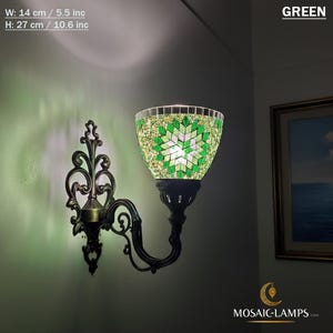 Può includere: Una lampada da parete con paralume a mosaico verde, larga 14 cm e alta 27 cm. La lampada ha una staffa in metallo scuro e ornata. Il paralume presenta un disegno a stella. La parola "GREEN" è visibile.