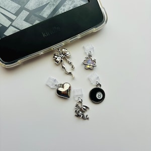 May include: An assortment of silver-toned charms, including a bow, heart, dragon, planet, and an eight ball, are displayed near a Kindle e-reader. Each charm is attached to a clear plastic clip, suggesting they are designed for use with the e-reader.