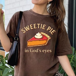 Op de afbeelding: Een bruin t-shirt met korte mouwen en een afbeelding van een stuk taart met slagroom. De tekst boven de taart luidt SWEETIE PIE en de tekst eronder luidt in God's eyes in een lichtgekleurd lettertype. Het shirt heeft een ronde hals.