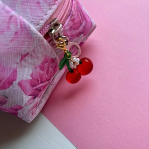 May include: A pink floral print cosmetic bag with a gold zip and a cherry charm. The charm features red cherries, green leaves, and small white flowers. The background is a solid pink.