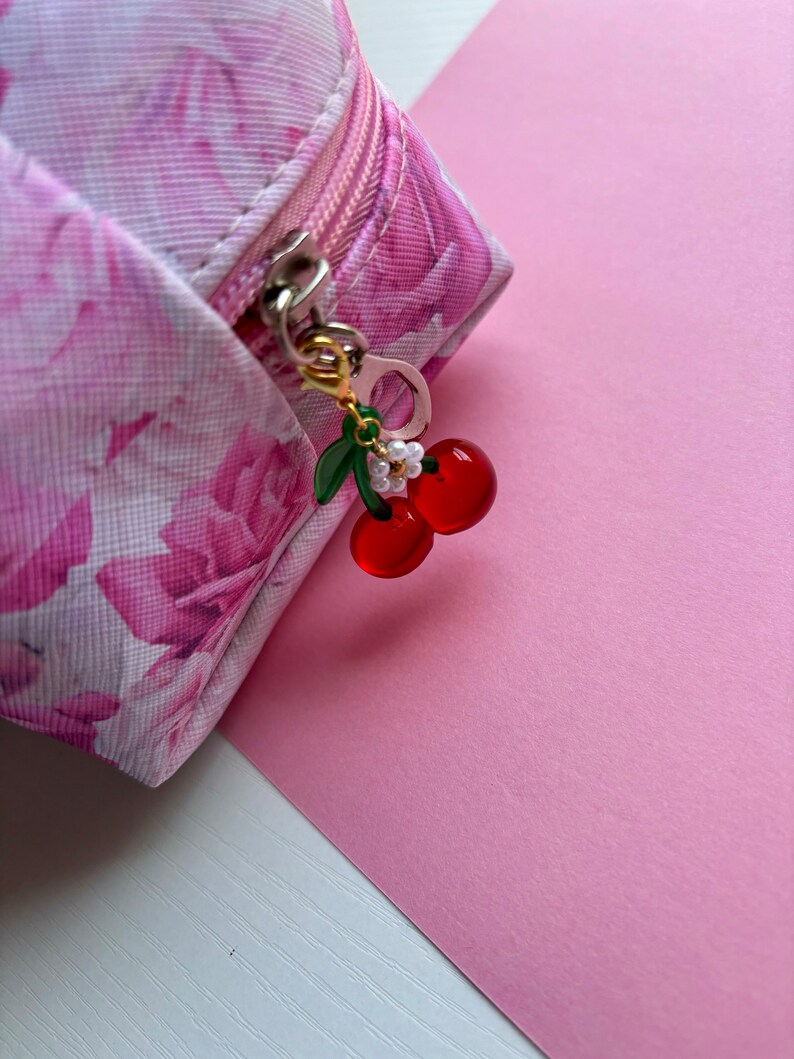 May include: A pink floral print cosmetic bag with a gold zip and a cherry charm. The charm features red cherries, green leaves, and small white flowers. The background is a solid pink.