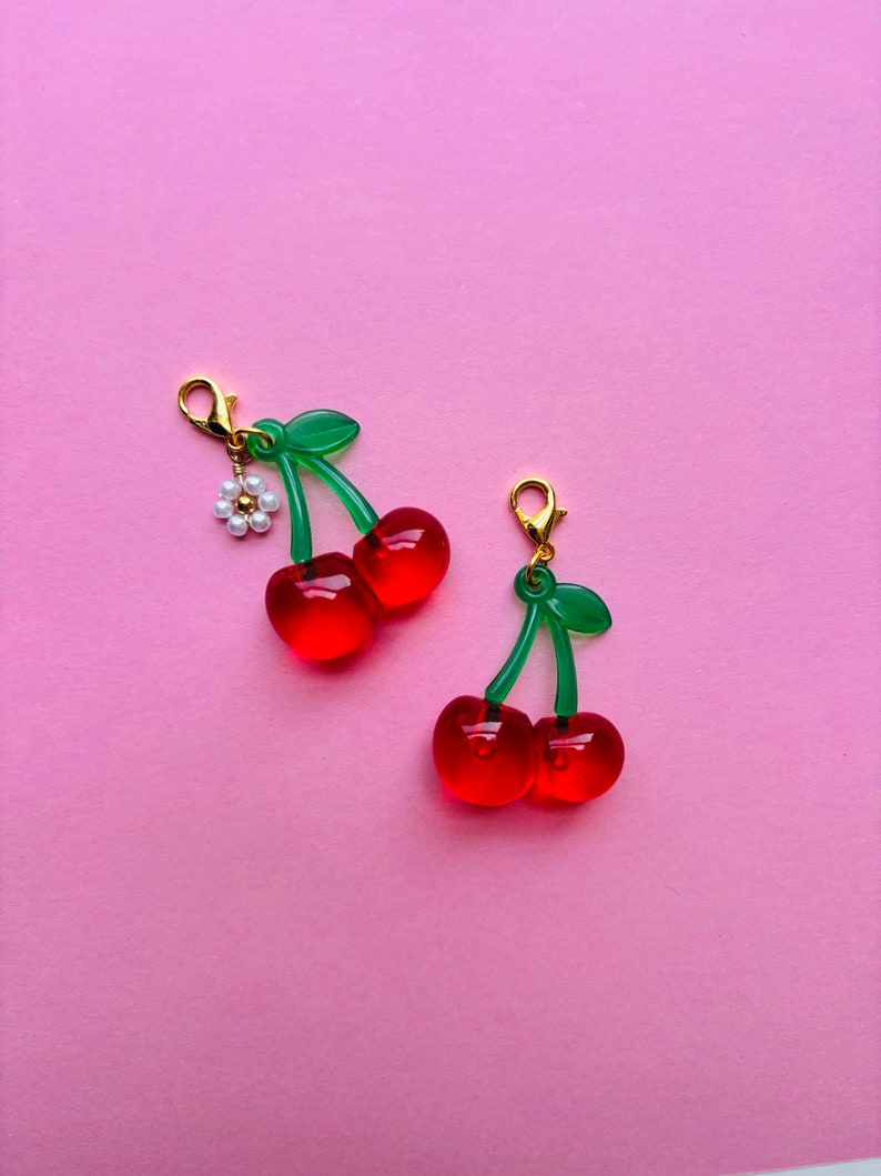 May include: Two cherry charms with red translucent fruit, green stems and leaves, and gold-coloured clasps. One charm has a small white flower with a pearl centre. The charms are set against a pink background.