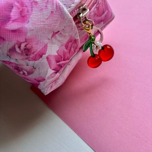 May include: A pink floral print cosmetic bag with a silver zip and a cherry charm. The charm features two red cherries, green leaves, and a small white flower. The bag is on a pink surface.