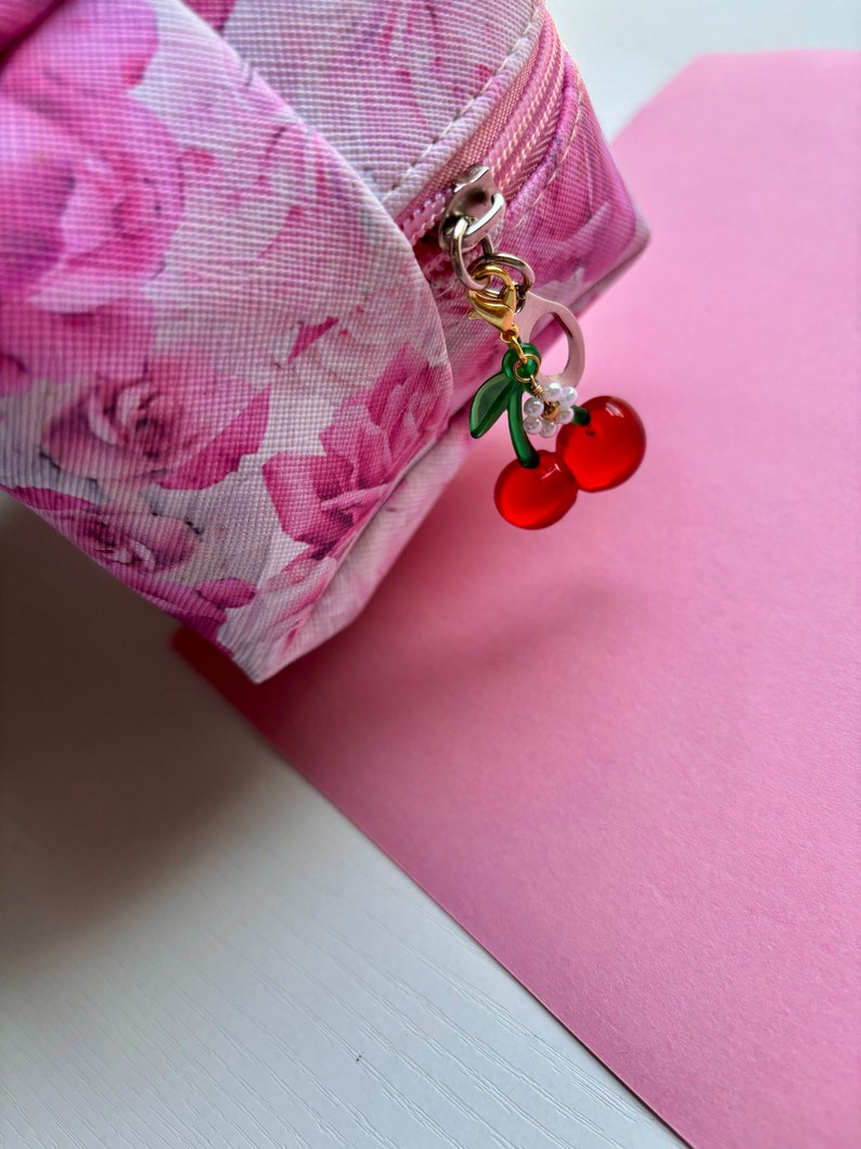 May include: A pink floral print cosmetic bag with a silver zip and a cherry charm. The charm features two red cherries, green leaves, and a small white flower. The bag is on a pink surface.