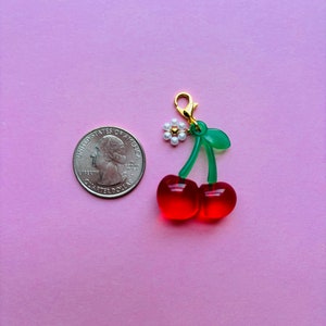 May include: A cherry charm with a gold clasp, green stem, and two red cherries. A small white flower with a gold center is attached to the stem. A U.S. quarter is next to the charm on a pink background.