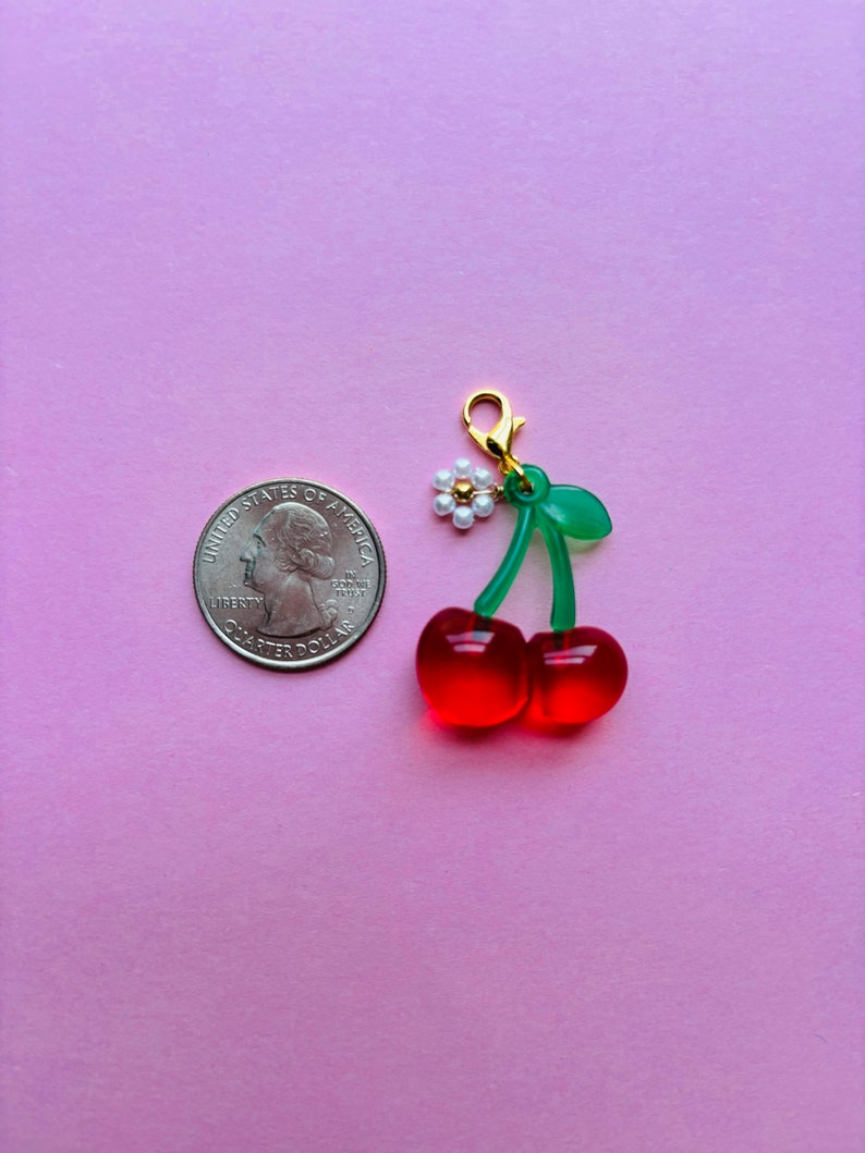 May include: A cherry charm with a gold clasp, green stem, and two red cherries. A small white flower with a gold center is attached to the stem. A U.S. quarter is next to the charm on a pink background.
