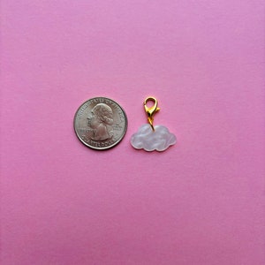 May include: A cloud-shaped charm with a gold-colored clasp. The charm is white with a pearlescent effect. A U.S. quarter is next to the charm for size comparison. The background is a solid pink color.