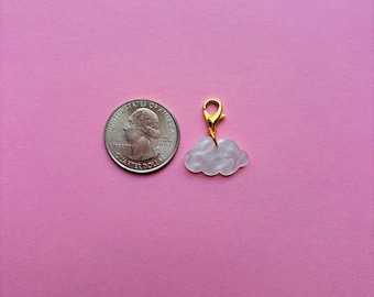 May include: A cloud-shaped charm with a gold-coloured clasp. The charm is white with a pearlescent effect. A U.S. quarter is next to the charm for size comparison. The background is a solid pink colour.