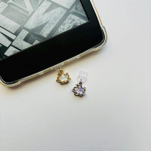 May include: Two small, celestial-themed charms. One is gold-toned with a white center, the other is silver-toned with an iridescent center. Both have a planet and star design. The charms are attached to a clear plastic piece, likely for a phone or e-reader.