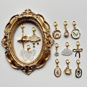 Puede incluir: Una colección de dijes de metal dorado con cierres de langosta, con formas que incluyen estrellas, nubes, mariposas, lunas, lazos y flores. Algunos dijes contienen resina translúcida, perlas o motivos florales. Se incluye un gran marco ovalado dorado ornamentado.