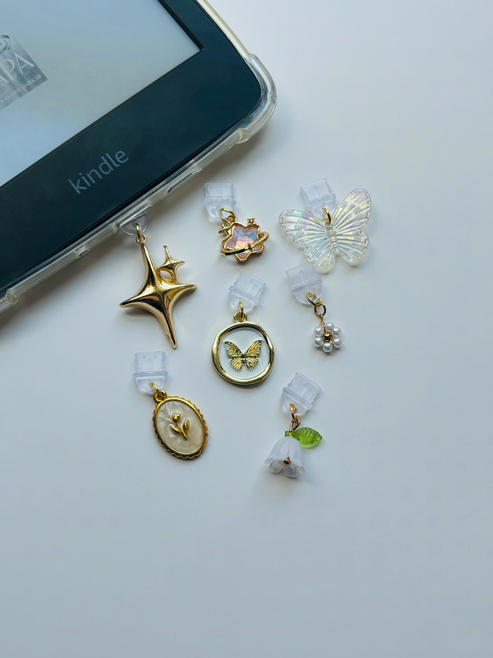A Kindle on a white surface beside eight decorative charms. Charms include butterflies, a star, a flower, and pearls.