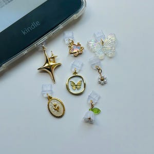 May include: A collection of decorative charms for a Kindle e-reader. The charms include a gold starburst, a gold and iridescent flower, a clear butterfly, a gold butterfly in a circle, a pearl cluster, a white flower, and a gold oval with a flower.