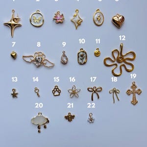 Puede incluir: Una colecci&oacute;n de dijes dorados en varias formas, incluyendo estrellas, mariposas, corazones, flores y lazos. Algunos dijes presentan detalles de cristal o esmalte. Los dijes est&aacute;n dispuestos sobre una superficie clara.