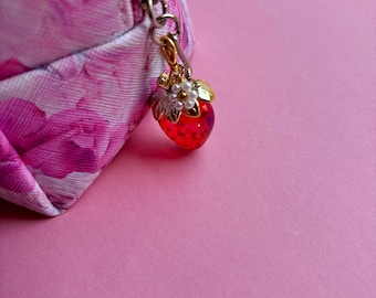 May include: A pink floral cosmetic bag with a silver zip and a strawberry charm. The charm is red with gold and white details. The background is a plain pink surface. The bag is partially visible.