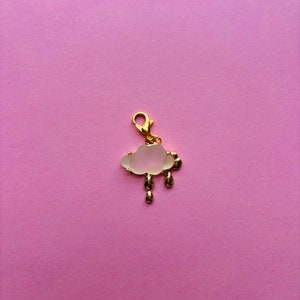 May include: A gold-colored charm in the shape of a cloud with three gold-colored raindrops. The cloud is white and has a gold-colored border. The charm has a gold-colored clasp and is set against a pink background.