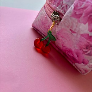 May include: A pink floral print cosmetic bag with a gold zip and a cherry charm. The charm features two red cherries and green leaves. The bag is on a pink surface.