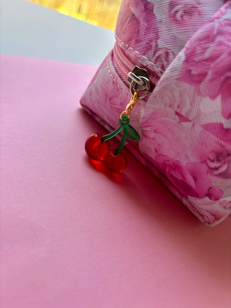 May include: A pink floral print cosmetic bag with a gold zip and a cherry charm. The charm features two red cherries and green leaves. The bag is on a pink surface.