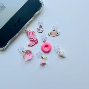 May include: A Kindle e-reader with a clear case is shown with several colorful charms attached. The charms include a pink boot, a cloud, a pink donut, a peach, a rose, a mushroom, and a pink planet. The word "Kindle" is visible on the device.