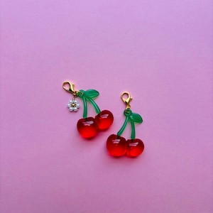 May include: Two pairs of cherry-shaped charms. Each charm features two translucent red cherries with green stems and leaves. One charm has a small white flower with gold accents. The charms have gold-coloured clasps.