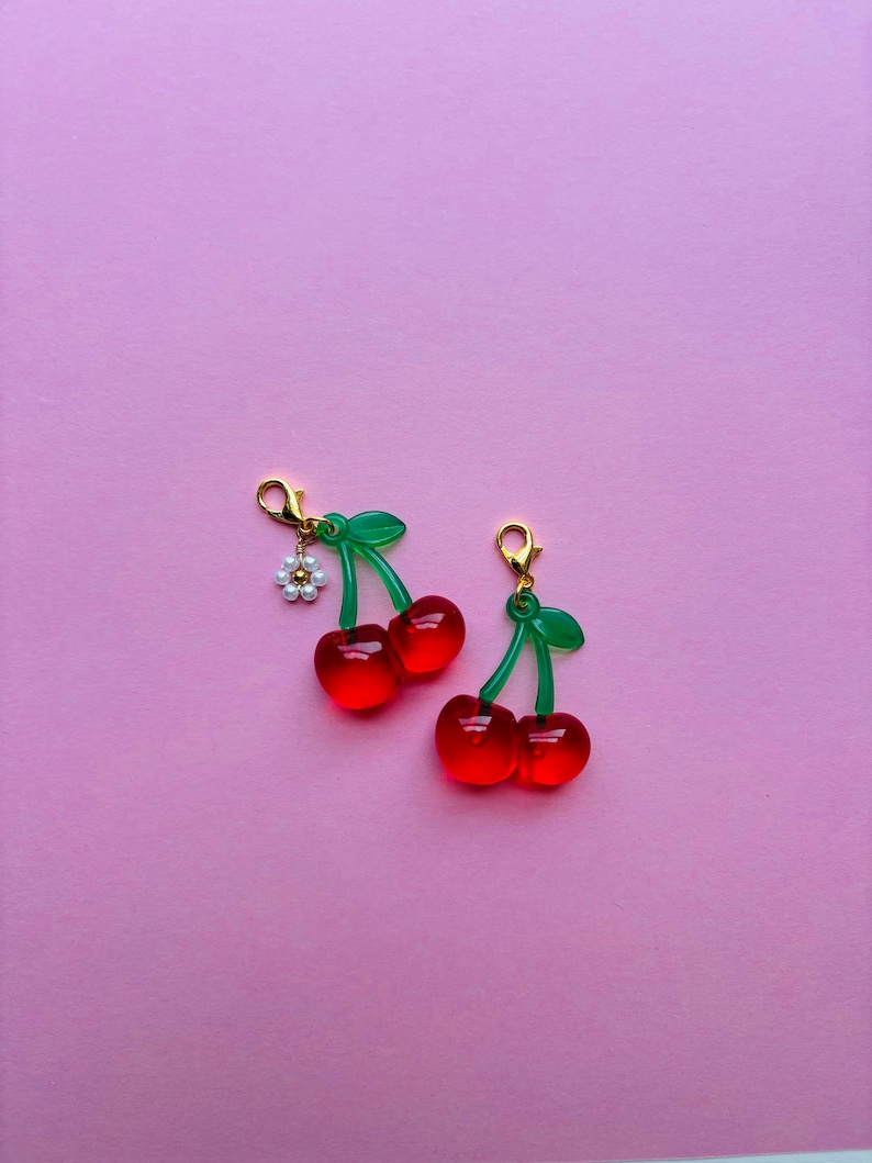 May include: Two pairs of cherry-shaped charms. Each charm features two translucent red cherries with green stems and leaves. One charm has a small white flower with gold accents. The charms have gold-coloured clasps.