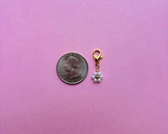 May include: A gold-coloured charm with a small daisy design, featuring white pearl-like beads and a gold centre. The charm is attached to a gold clasp. A U.S. quarter coin is next to the charm on a pink background.