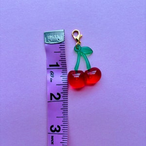 May include: A cherry charm with a gold-coloured clasp. The charm features two red cherries with green stems and leaves. A purple measuring tape is next to the charm, showing a length of approximately 3.8 cm.