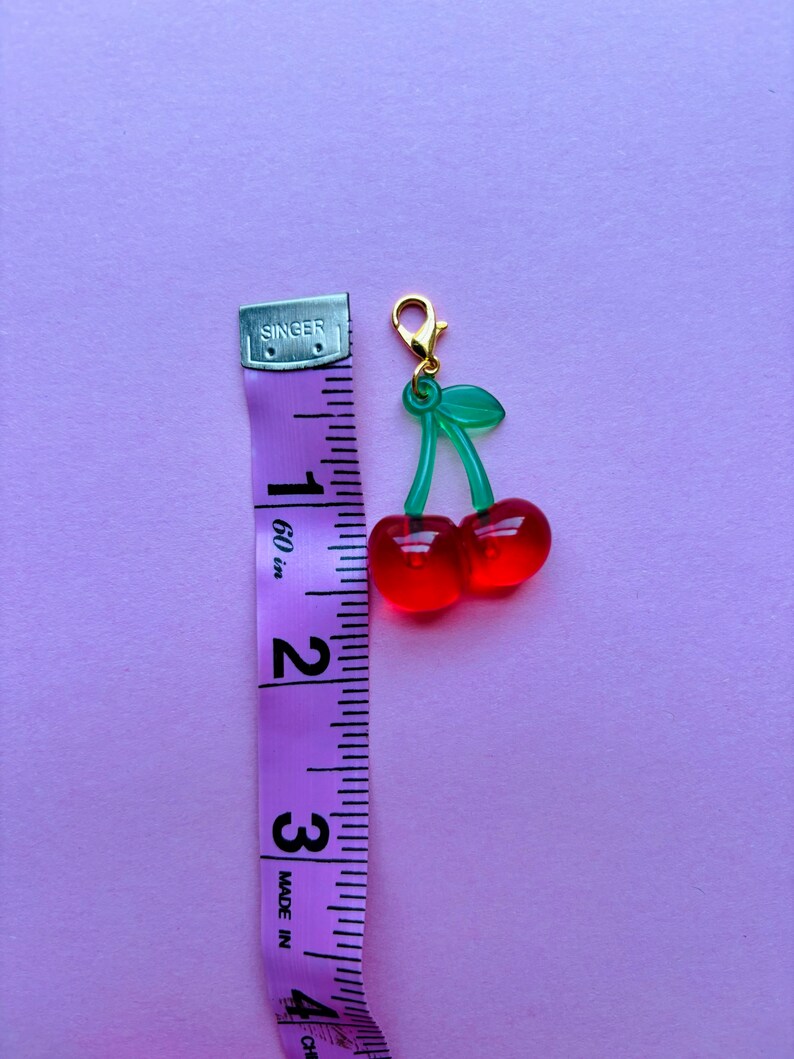 May include: A cherry charm with a gold-coloured clasp. The charm features two red cherries with green stems and leaves. A purple measuring tape is next to the charm, showing a length of approximately 3.8 cm.