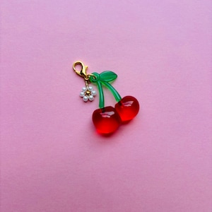 May include: A cherry charm with a gold clasp, green stems, and two red translucent cherries. A small white flower with pearl-like beads hangs from the clasp. The charm is set against a pink background.