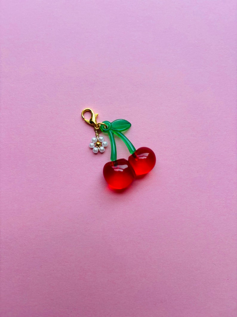 May include: A cherry charm with a gold clasp, green stems, and two red translucent cherries. A small white flower with pearl-like beads hangs from the clasp. The charm is set against a pink background.