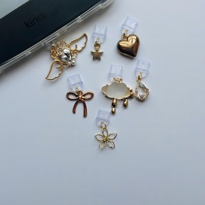 Puede incluir: Una colecci&oacute;n de colgantes dorados para tel&eacute;fono, que incluyen un coraz&oacute;n, una estrella, un lazo, una nube, una flor y un dije con alas. Los dijes est&aacute;n unidos a clips de pl&aacute;stico transparente, para una funda de tel&eacute;fono.