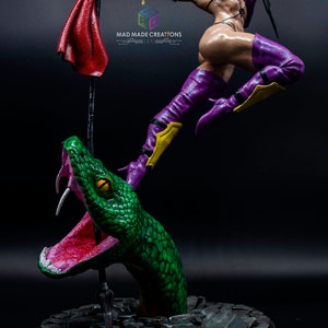 May include: A purple and yellow action figure of a female warrior standing on a green snake with its mouth open. The figure is holding a red cloth and a sword. The base of the figure is a gray stone circle.