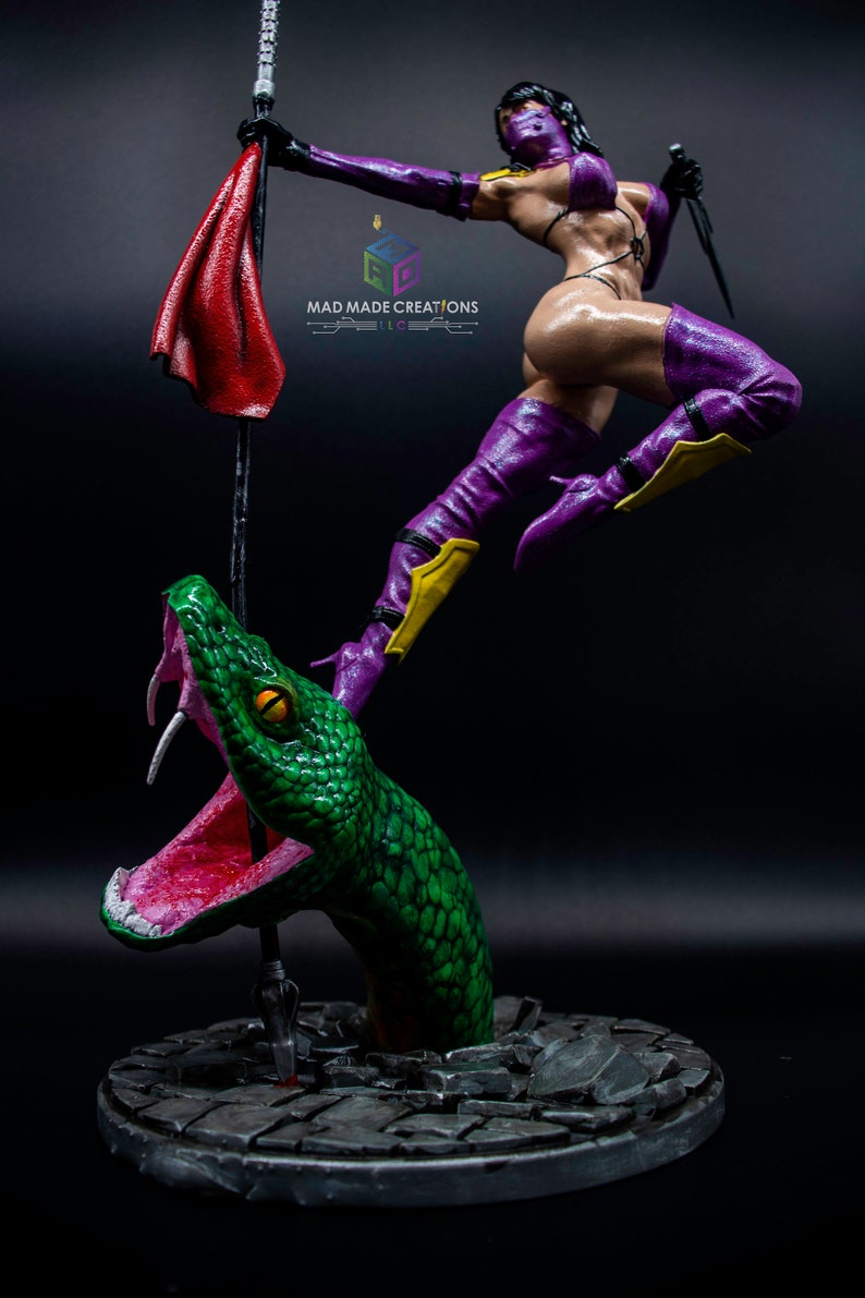 May include: A purple and yellow action figure of a female warrior standing on a green snake with its mouth open. The figure is holding a red cloth and a sword. The base of the figure is a gray stone circle.