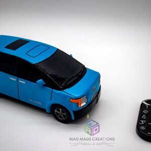 May include: A blue toy car with a black roof and a sunroof. The car has the word "PRANCER" printed on the side. The car is next to a black remote control with buttons.