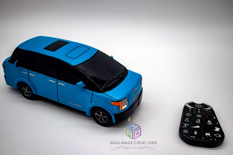 May include: A blue toy car with a black roof and a sunroof. The car has the word "PRANCER" printed on the side. The car is next to a black remote control with buttons.
