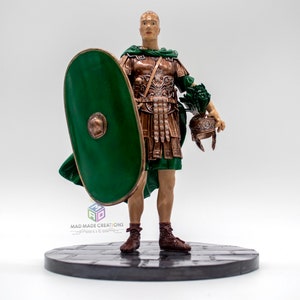 May include: A hand-painted figurine of a Roman soldier wearing a green tunic and bronze armor, holding a green shield and a helmet. The figurine is standing on a black base with a stone-like texture.