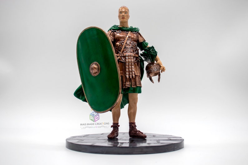 May include: A hand-painted figurine of a Roman soldier wearing a green tunic and bronze armor, holding a green shield and a helmet. The figurine is standing on a black base with a stone-like texture.