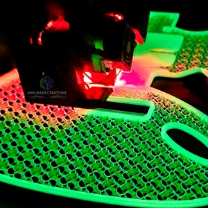 May include: A 3D printer is printing a green and red guitar body with a honeycomb pattern. The printer head is moving across the surface of the guitar body, depositing plastic filament.