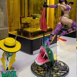 May include: Three 3D printed figurines: a green snake with its mouth open, a yellow-hatted masked figure, and a female figure in a purple outfit holding a spear. The figures are on a white surface. The female figure is standing on a base with a cobblestone design. The snake is on a similar base. The masked figure is on a small base with the word "MASK" printed on it.