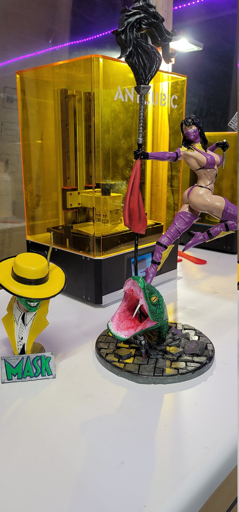 May include: Three 3D printed figurines: a green snake with its mouth open, a yellow-hatted masked figure, and a female figure in a purple outfit holding a spear. The figures are on a white surface. The female figure is standing on a base with a cobblestone design. The snake is on a similar base. The masked figure is on a small base with the word "MASK" printed on it.