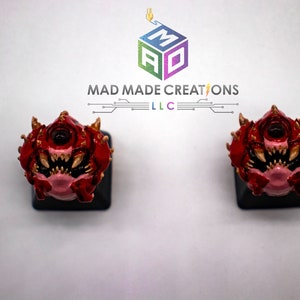 Puede incluir: Dos teclas de teclado impresas en 3D de color rojo y dorado con un solo ojo y dientes afilados. Las teclas están sobre una base negra y se colocan sobre un fondo blanco. El texto "MAD MADE CREATIONS LLC" es visible sobre las teclas.